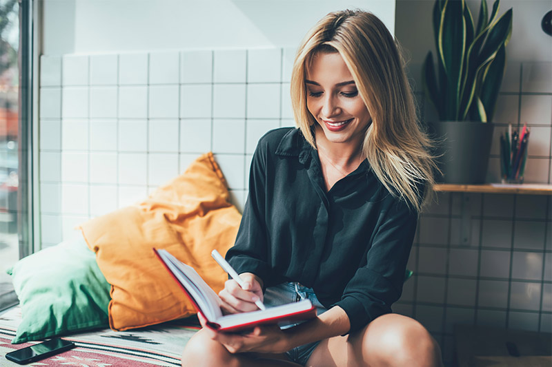 A smiling woman with long blonde hair sits indoors on a cushioned bench, writing in a red notebook. She wears a black shirt and denim shorts. There are colorful pillows and a plant in the background.